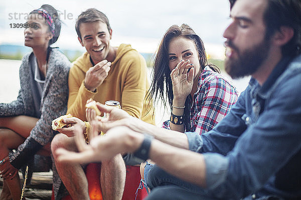 Glückliche Freunde halten Hotdogs  während sie am Fluss sitzen