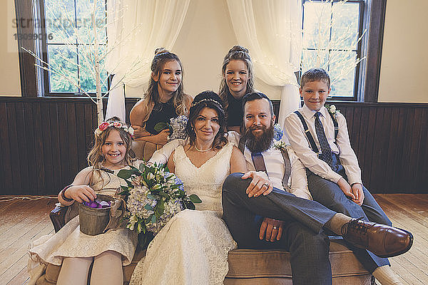 Porträt eines frisch vermählten Paares  das während der Hochzeitszeremonie von Pagen und Blumenmädchen mit den Brautjungfern auf dem Sofa sitzt