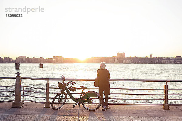 Rückansicht einer älteren Frau  die bei Sonnenuntergang am Fahrrad gegen das Meer steht