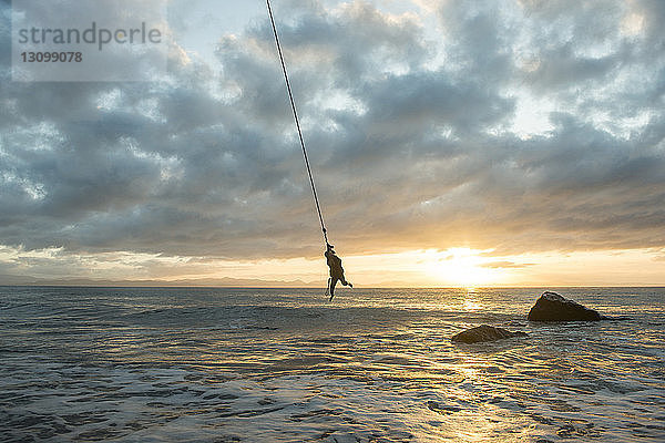 Silhouetten-Mann hängt am Seil über dem Meer vor bewölktem Himmel bei Sonnenuntergang