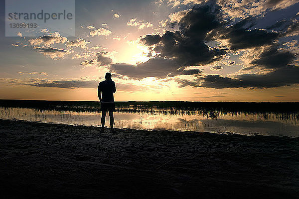 Silhouettenmann steht am Seeufer vor bewölktem Himmel bei Sonnenuntergang