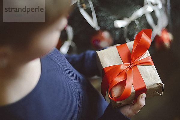 Nahaufnahme eines Jungen  der zu Hause ein Geschenk am Weihnachtsbaum hält