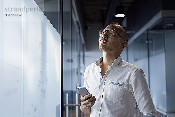 Nachdenklicher Geschäftsmann hält Smartphone in der Hand  während er an der Glaswand im Büro steht