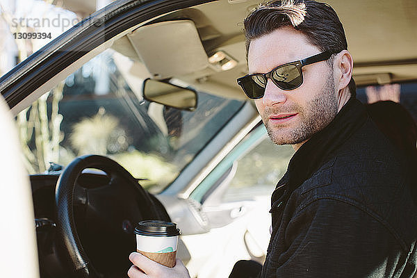 Hübscher Mann mit Sonnenbrille  der einen Einwegbecher hält  während er im Auto sitzt