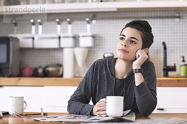 Frau mit Zeitungen und Kaffeetasse schaut weg  während sie am Esstisch sitzt