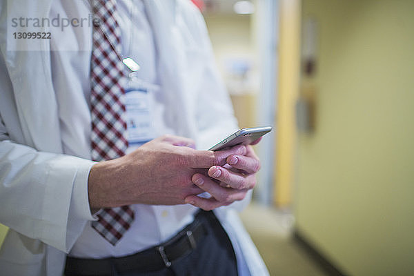 Männlicher Arzt benutzt Mobiltelefon  während er im Bürokorridor steht