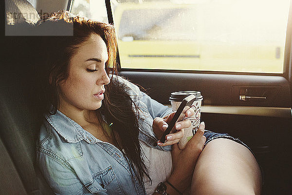 Frau mit Einwegbecher  die während der Fahrt im Auto ein Mobiltelefon benutzt