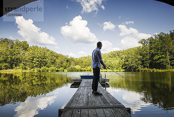 Mann in voller Länge angelt  während er auf einem Holzsteg über dem See vor Bäumen und bewölktem Himmel steht