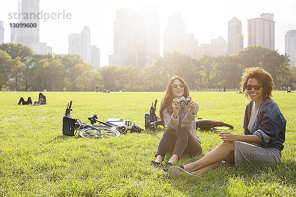 Glückliche Frau hält Kamera und entspannt sich mit Freundin auf Grasfeld