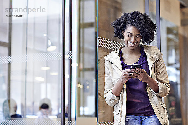 Glückliche Geschäftsfrau benutzt Smartphone  während sie im Büro an der Tür steht
