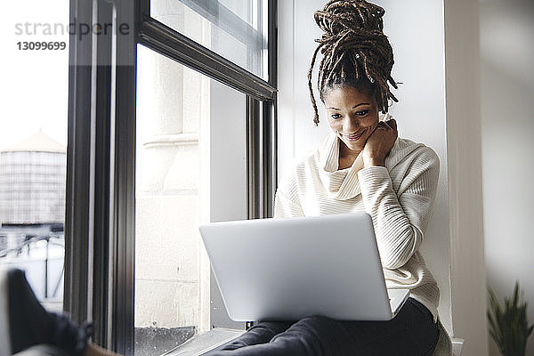 Lächelnde Geschäftsfrau benutzt Laptop  während sie im Büro am Fensterbrett sitzt