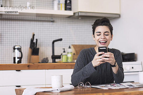 Glückliche Frau benutzt Mobiltelefon am Esstisch in der Küche