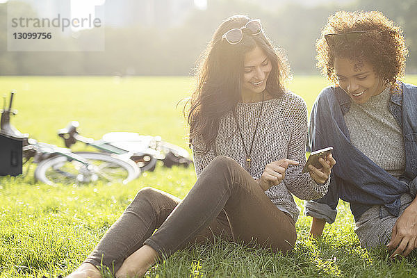 Freunde benutzen Mobiltelefon  während sie auf einem Grasfeld sitzen