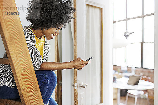 Glückliche Frau benutzt Mobiltelefon  während sie zu Hause auf der Leiter sitzt