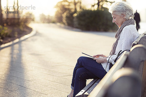 Ältere Frau benutzt einen Tablet-Computer  während sie auf einer Bank am Fußweg sitzt