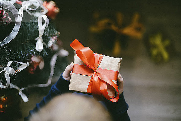 Abgetrennte Hände eines Jungen  der zu Hause ein Geschenk am Weihnachtsbaum hält