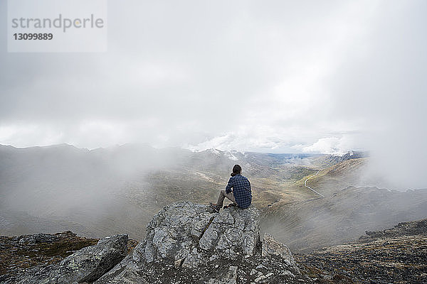 Rückansicht eines Mannes  der bei nebligem Wetter auf einer Klippe vor der Landschaft sitzt