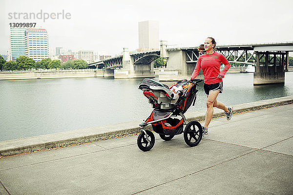 Frau joggt mit Kinderwagen auf Fußweg am Fluss gegen Brücke