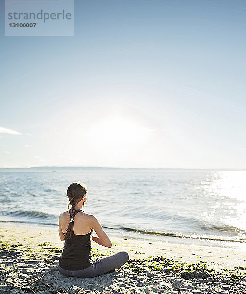 Rückansicht einer am Strand meditierenden Frau  die am sonnigen Tag gegen den Himmel meditiert