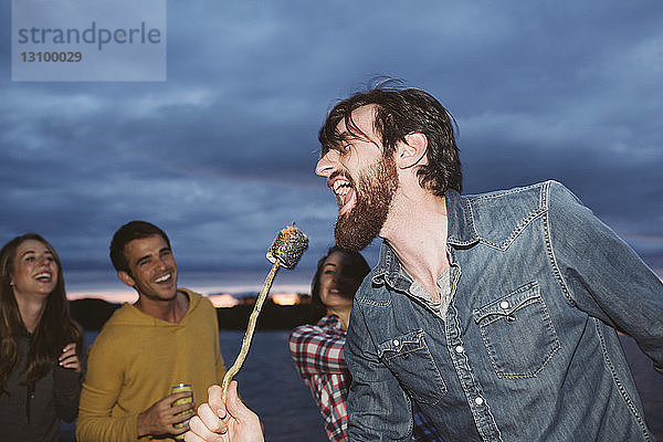 Lächelnde Freunde schauen den Mann singend an  während sie gebratenen Marshmallow am Stock gegen den bewölkten Himmel halten