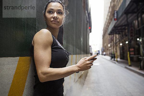 Nachdenkliche sportliche Frau  die ein Smartphone in der Hand hält  während sie sich an eine Wand an der Straße lehnt