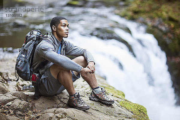 Nachdenklicher männlicher Wanderer sitzt auf einem Felsen am Wasserfall
