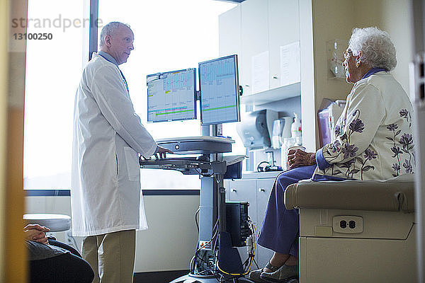 Männlicher Arzt spricht mit weiblicher Patientin  während er den Computer auf der Krankenhausstation benutzt