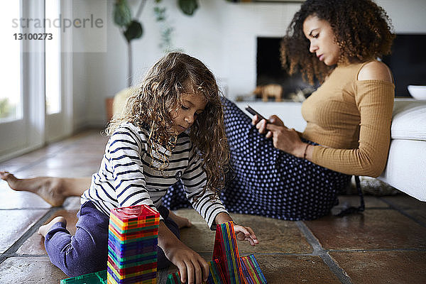 Mutter benutzt Mobiltelefon  während die sitzende Tochter im Wohnzimmer mit Spielzeug spielt