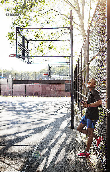Mann hält Mobiltelefon in der Hand  während er sich am Zaun des Basketballplatzes anlehnt