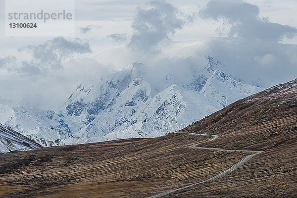 Landschaftliche Ansicht der Straße inmitten des Feldes vor schneebedeckten Bergen im Denali-Nationalpark und -Schutzgebiet bei nebligem Wetter