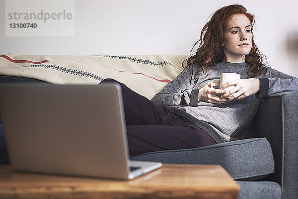 Nachdenkliche Frau mit Kaffeetasse  die beim Entspannen auf dem Sofa wegschaut