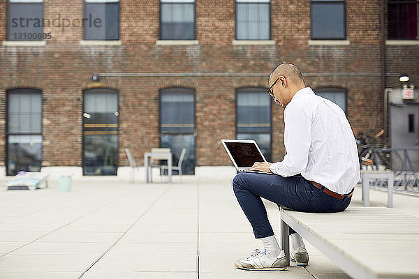 Geschäftsmann in voller Länge mit Laptop-Computer  während er auf einem Liegestuhl auf der Gebäudeterrasse sitzt