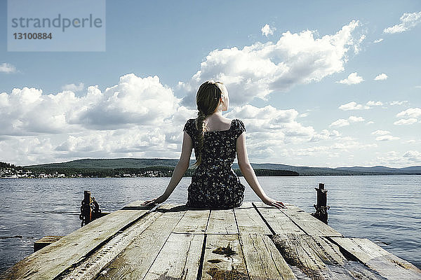 Nachdenkliche Frau sitzt am Pier über dem Fluss vor bewölktem Himmel