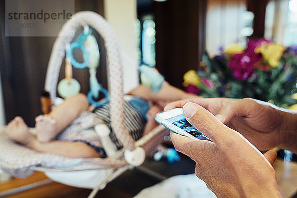 Ausgeschnittenes Bild einer Hand mit einem Smartphone von einem Baby im Kindersitz zu Hause