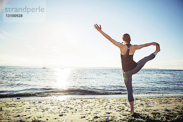 Rückansicht einer Frau  die an einem sonnigen Tag am Strand eine Yogastellung von der Hand bis zum großen Zeh praktiziert