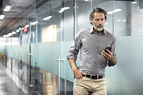 Geschäftsmann mit Hand in der Tasche  der in der Büro-Lobby auf ein Smartphone schaut