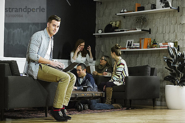 Porträt eines Geschäftsmannes  der einen Laptop benutzt  während sich Kollegen im Hintergrund im Büro unterhalten