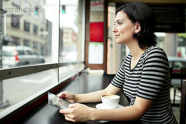 Nachdenkliche Frau hält Tablet-Computer in der Hand  während sie durch ein Café-Fenster schaut