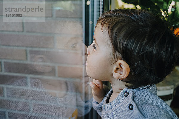 Seitenansicht eines kleinen Mädchens  das Glas küsst  während es zu Hause durch ein Fenster schaut