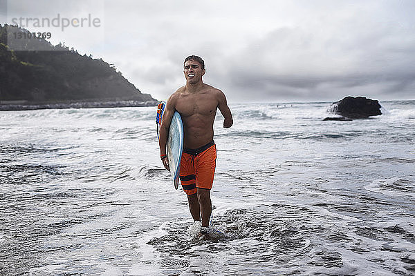 Behinderter Mann trägt Surfbrett  während er aus dem Meer gegen den Himmel läuft