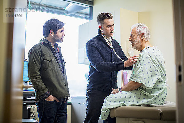 Männliche Ärzte untersuchen Patient  der auf der Krankenhausstation auf dem Bett sitzt und durch die Tür gesehen wird