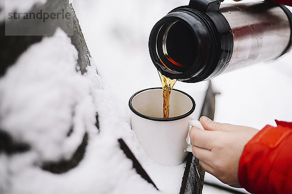 Abgehackte Hände einer Frau  die schwarzen Kaffee aus isoliertem Getränkebehälter in Becher auf Schnee schüttet