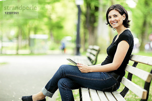 Porträt einer glücklichen Frau  die einen Tablet-Computer hält  während sie auf einer Parkbank sitzt