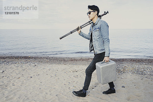 Junger Mann in voller Länge mit Aktentasche und Stativ beim Strandspaziergang