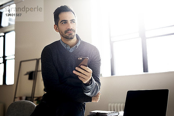 Nachdenklicher Geschäftsmann  der ein Smartphone in der Hand hält und im Büro sitzt