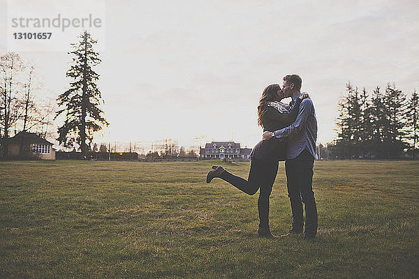 Romantisches Paar küsst sich im Stehen auf Grasfeld gegen den Himmel