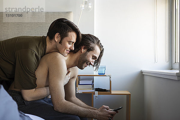 Mann umarmt Freund mit Mobiltelefon  während er auf dem Bett sitzt