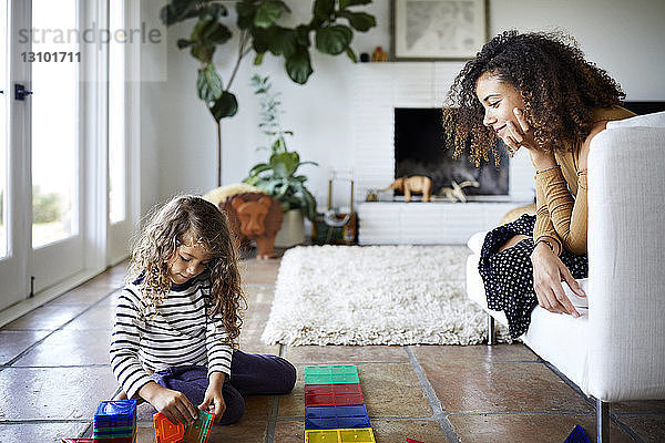 Mutter betrachtet Tochter beim Spielen mit Spielzeugklötzen im Wohnzimmer