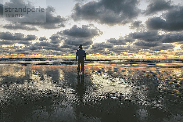 Rückansicht eines Wanderers  der bei Sonnenuntergang am Long Beach vor bewölktem Himmel steht