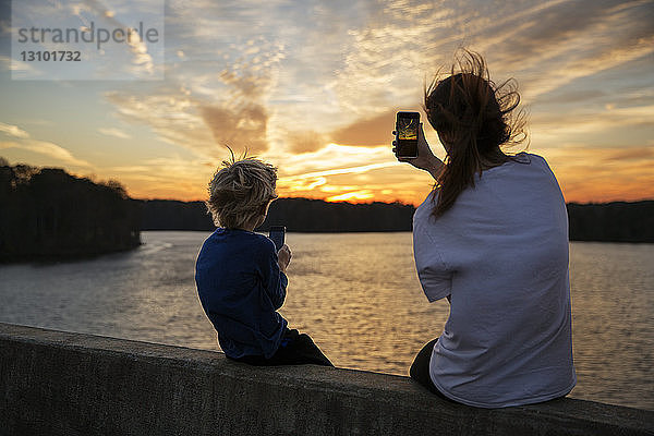 Junge und Mädchen sitzen auf einem Felsvorsprung und fotografieren den Sonnenuntergang